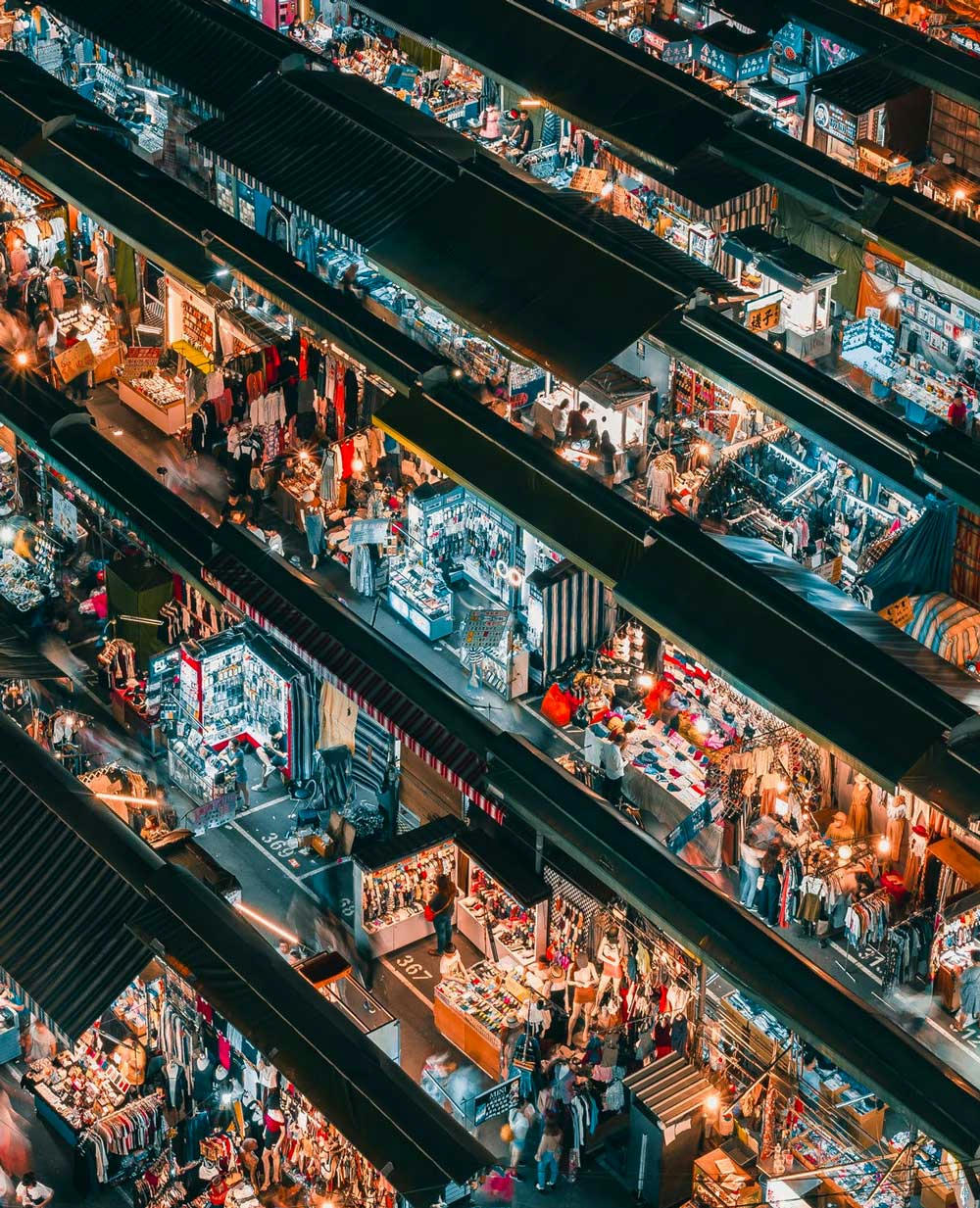 Flygbild av en livlig nattmarknad med rader av marknadsstånd som säljer olika varor, belyst av färgglada ljus i en tät och välorganiserad layout.