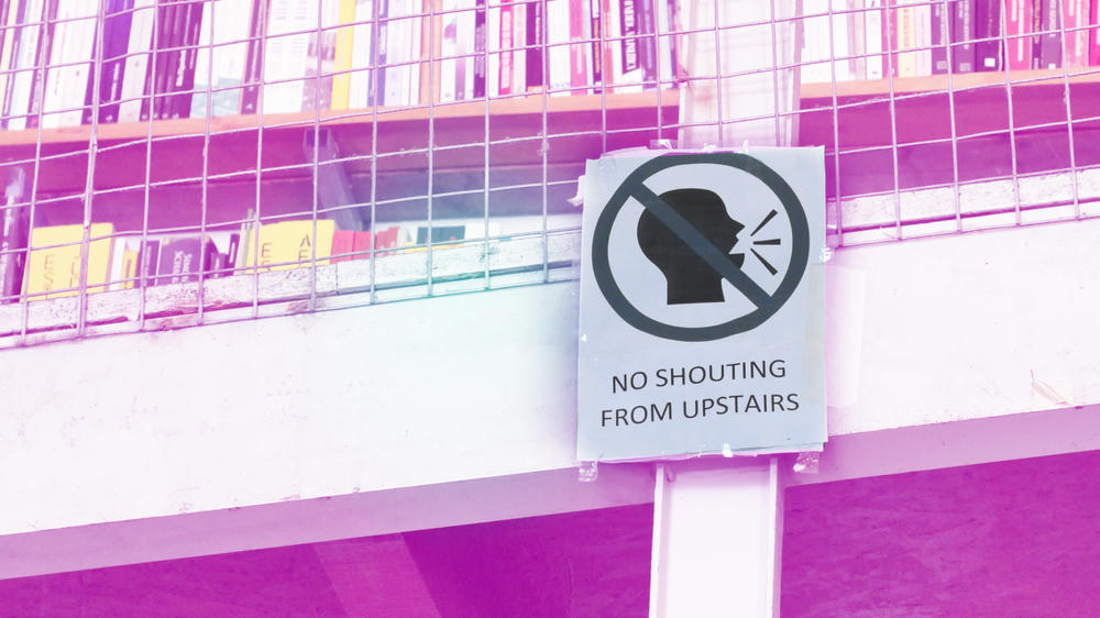 Skylt med texten ‘No shouting from upstairs’ och en symbol som förbjuder ropande, uppsatt på ett räcke med böcker i bakgrunden.