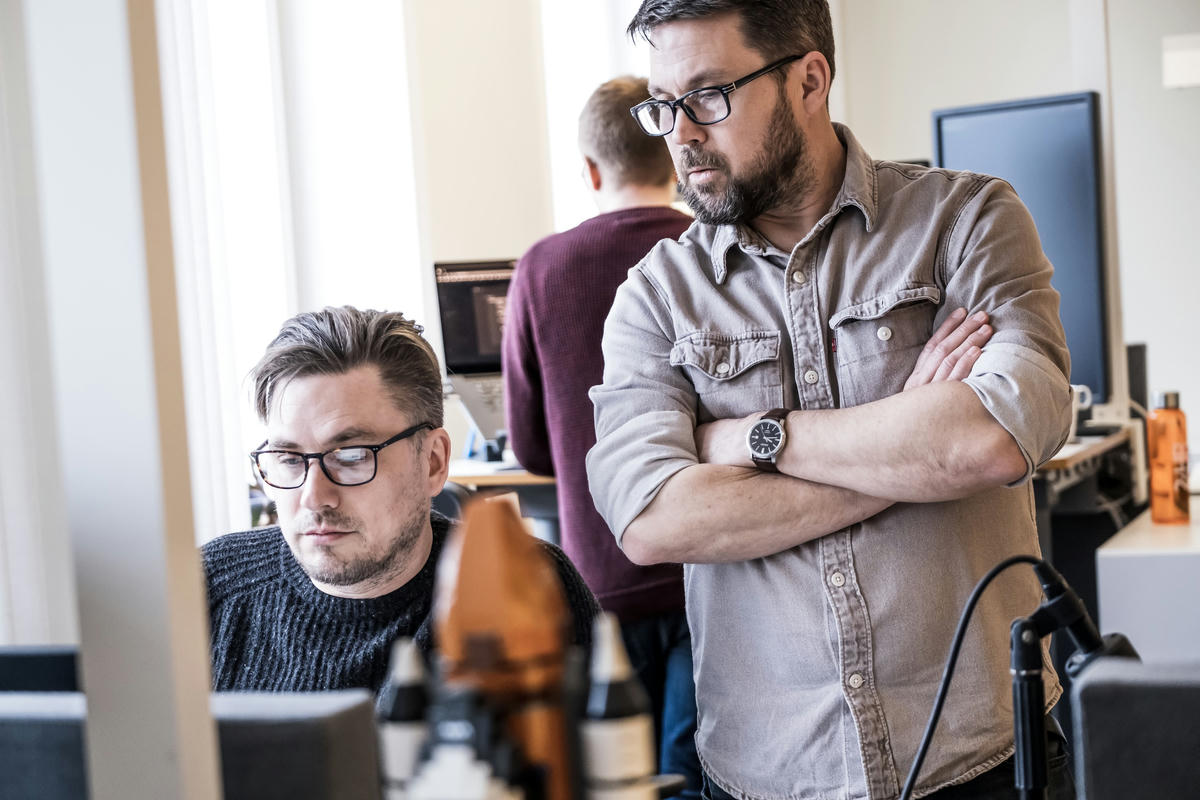 Två kollegor på TwoFour diskuterar och samarbetar vid sina datorer på kontoret. En av dem står och tittar på skärmen medan den andra sitter vid skrivbordet.