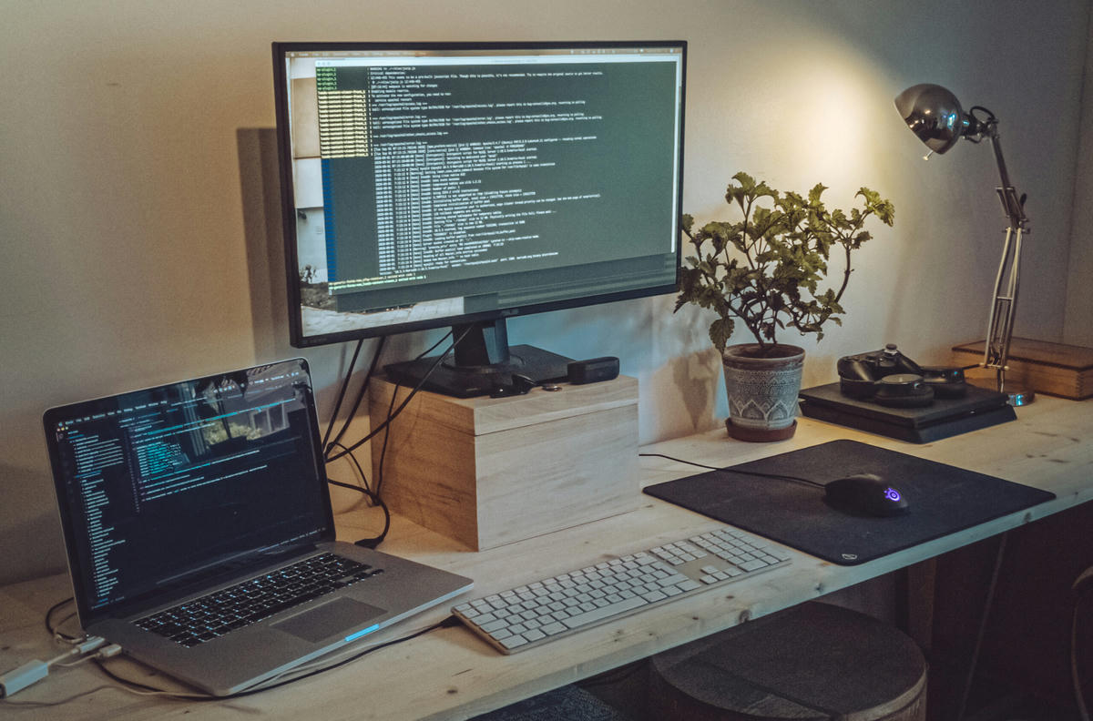 En arbetsstation med en bärbar dator, en extern bildskärm som visar kod, ett tangentbord, en mus, en skrivbordslampa och en krukväxt på ett träbord. En arbetsstation med en bärbar dator, en extern bildskärm som visar kod, ett tangentbord, en mus, en skrivbordslampa och en krukväxt på ett träbord.