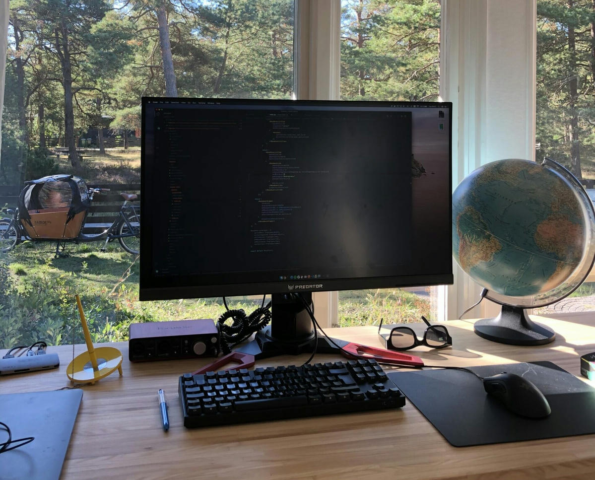Arbetsstation med en extern bildskärm som visar kod, ett tangentbord, en mus, en jordglob och andra skrivbordstillbehör. Stora fönster i bakgrunden med utsikt över en trädgård och en lådcykel. Arbetsstation med en extern bildskärm som visar kod, ett tangentbord, en mus, en jordglob och andra skrivbordstillbehör. Stora fönster i bakgrunden med utsikt över en trädgård och en lådcykel.
