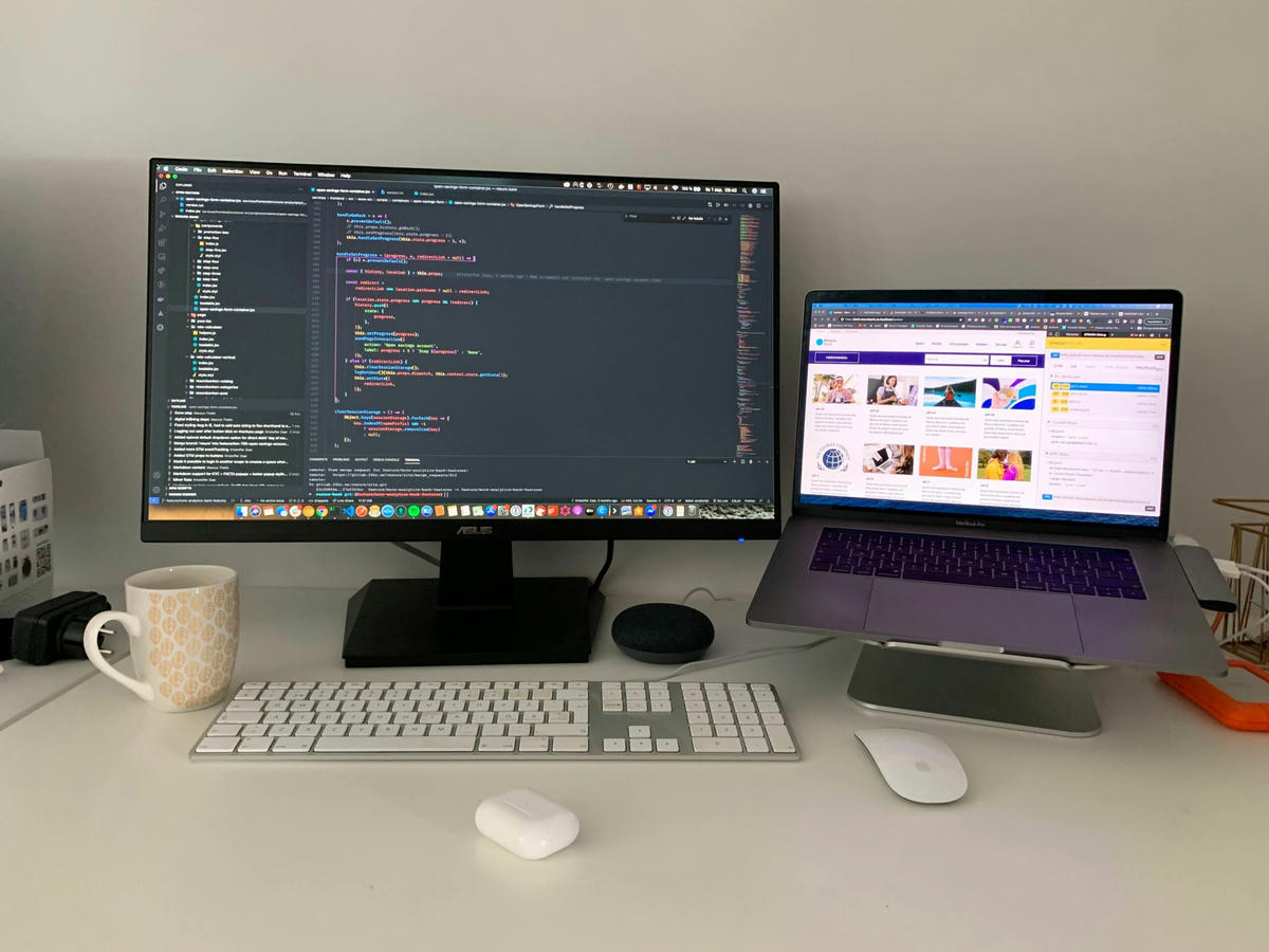 Arbetsstation med en extern bildskärm som visar kod, en bärbar dator på ett stativ som visar en webbsida, ett tangentbord, en mus, en kaffemugg och ett par trådlösa hörlurar på ett vitt skrivbord. Arbetsstation med en extern bildskärm som visar kod, en bärbar dator på ett stativ som visar en webbsida, ett tangentbord, en mus, en kaffemugg och ett par trådlösa hörlurar på ett vitt skrivbord.