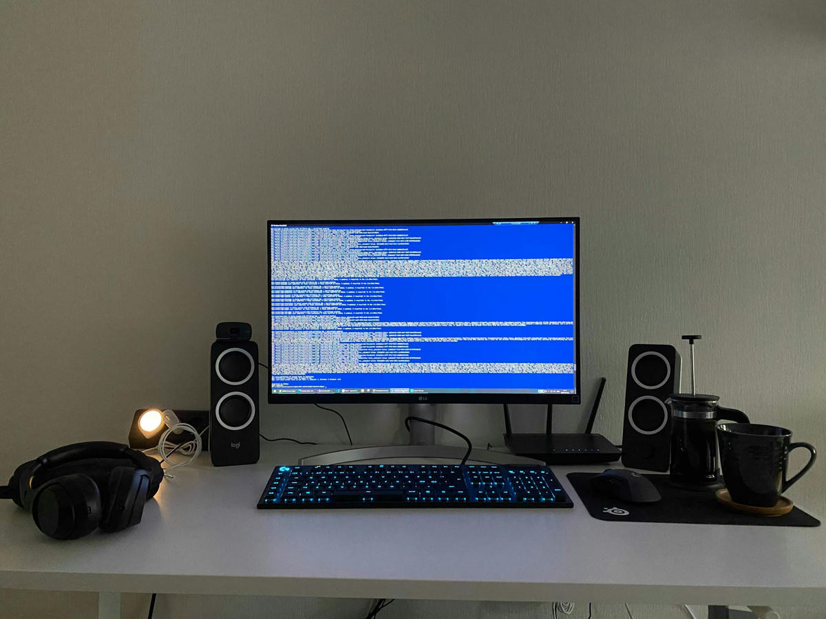 Arbetsstation med en extern bildskärm som visar kod, ett mekaniskt tangentbord med blå bakgrundsbelysning, en mus, högtalare, hörlurar, en bordslampa och en kaffemugg med en kaffepress på ett vitt skrivbord. Arbetsstation med en extern bildskärm som visar kod, ett mekaniskt tangentbord med blå bakgrundsbelysning, en mus, högtalare, hörlurar, en bordslampa och en kaffemugg med en kaffepress på ett vitt skrivbord.