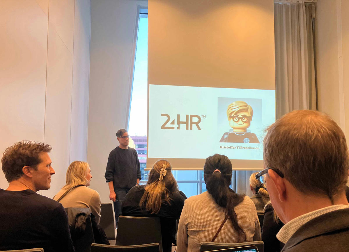 Föreläsning eller workshop med TwoFour: Kristoffer Yi Fredriksson presenterar inför en publik med en bild av en LEGO-figur och TwoFour-logotypen på projektorduken i bakgrunden. Föreläsning eller workshop med TwoFour: Kristoffer Yi Fredriksson presenterar inför en publik med en bild av en LEGO-figur och TwoFour-logotypen på projektorduken i bakgrunden.