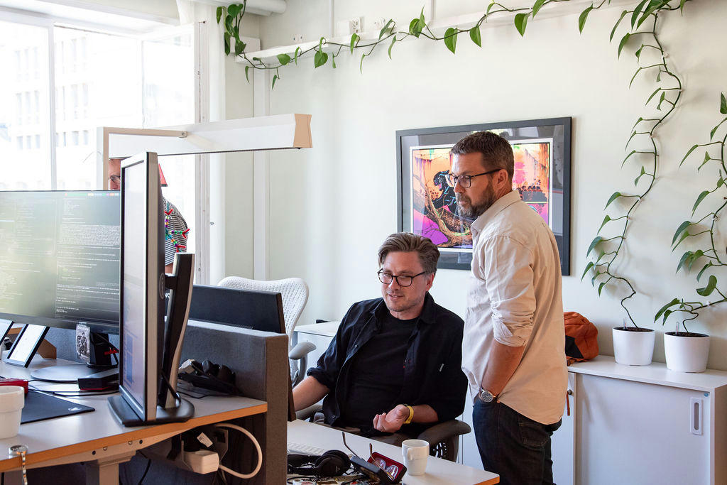 Två kollegor på TwoFour diskuterar och samarbetar vid sina datorer på kontoret. En av dem står och tittar på skärmen medan den andra sitter vid skrivbordet. Två kollegor på TwoFour diskuterar och samarbetar vid sina datorer på kontoret. En av dem står och tittar på skärmen medan den andra sitter vid skrivbordet.