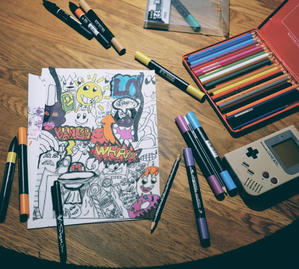 En teckning fylld med färgglada karaktärer och ord som ‘BANG!’ och ‘WHAM!’, omgiven av färgpennor, markers och en retro-spelenhet på ett träbord.